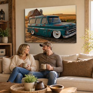 A large rectangular wall art print showing a vintage teal 1966 Chevy Suburban Carryall parked in a field at sunset, displayed above a beige sofa in a living room with a couple holding mugs.