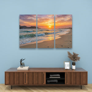 A three-panel canvas wall art print depicting a beach sunset with ocean waves, displayed on a light blue wall above a wooden media console with decorative items.
