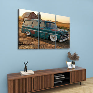A three-panel canvas wall art print depicting a vintage teal 1966 Chevy Suburban Carryall parked in a rural field at sunset, displayed above a wooden console cabinet in a styled room with a blue wall.