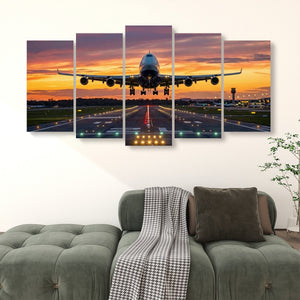 A 5-panel canvas wall art set depicting a commercial airplane landing on an airport runway during a vibrant sunset, displayed on a wall above a green sofa in a living room setting.