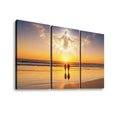 Three-panel canvas wall art showing a beach sunset scene with a glowing white figure above two silhouetted figures walking on the sand, displayed against a light gray background.