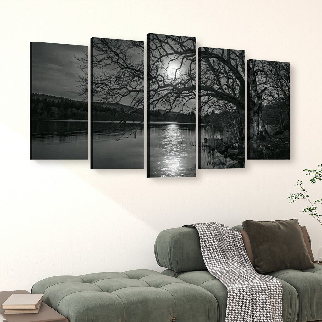 A five-panel black and white canvas wall art set depicting a moonlit lake scene with bare tree branches over the water, displayed above a green sofa in a living room setting.