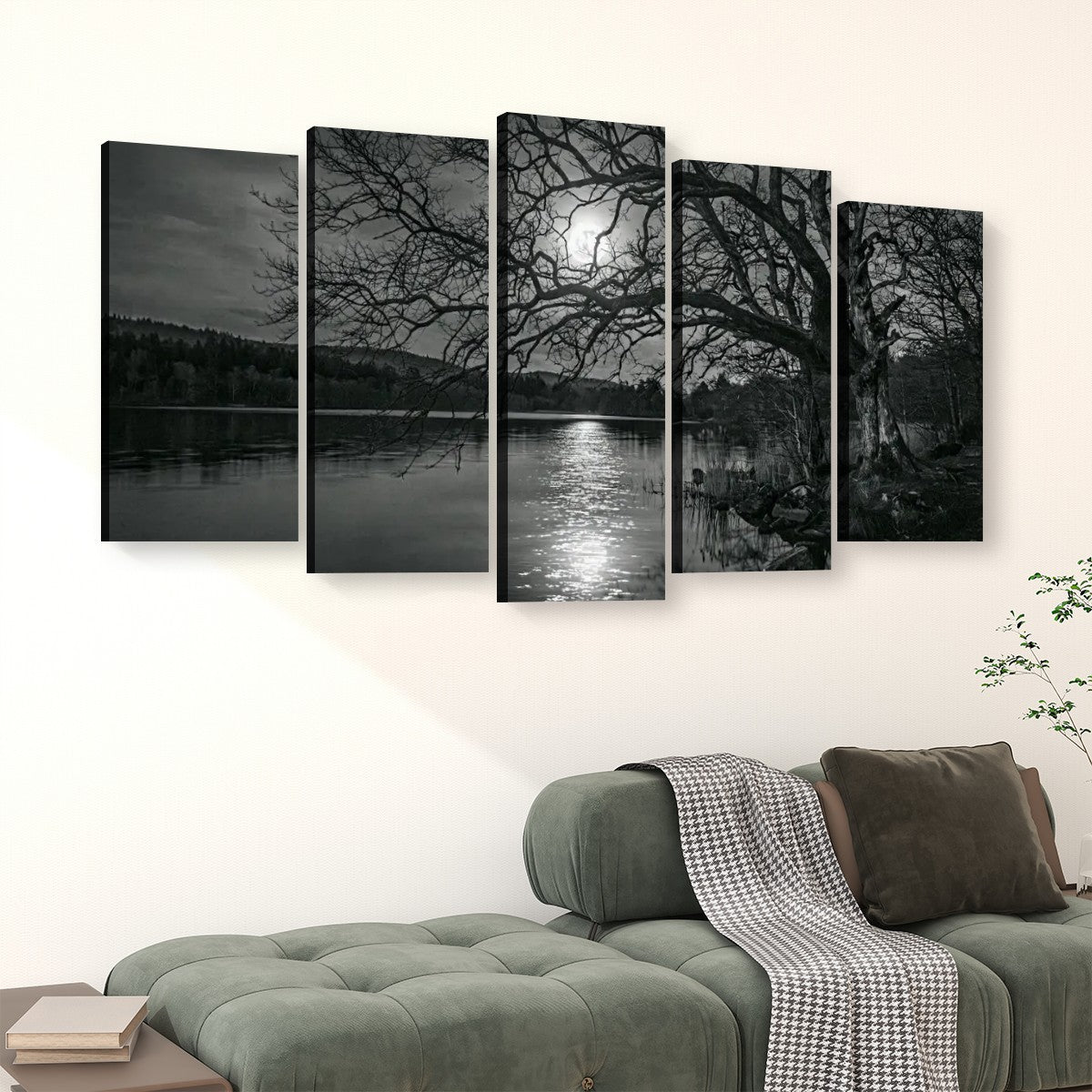 A five-panel black and white canvas wall art set depicting a moonlit lake scene with bare tree branches over the water, displayed above a green sofa in a living room setting.