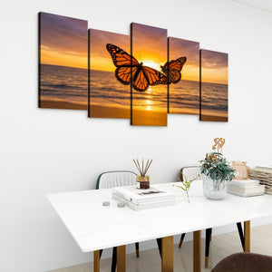 A five-panel canvas wall art set depicting a monarch butterfly over an ocean sunset, displayed on a white wall above a dining table in a modern room setting.