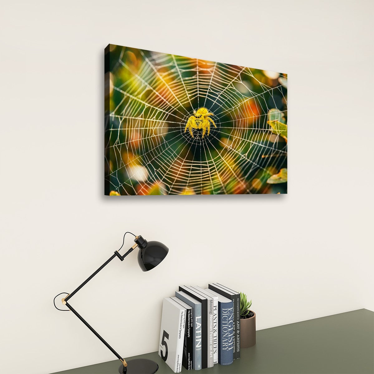 A rectangular wall art print showing a close-up spiderweb with a yellow spider in the center, displayed on a light wall above a desk with a black lamp and books.