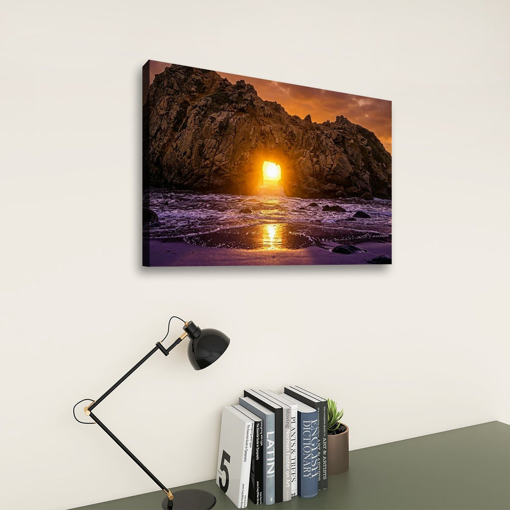 A rectangular wall art print showing a rocky coastline at sunset hangs on a light beige wall above a dark gray table with a black desk lamp and books.