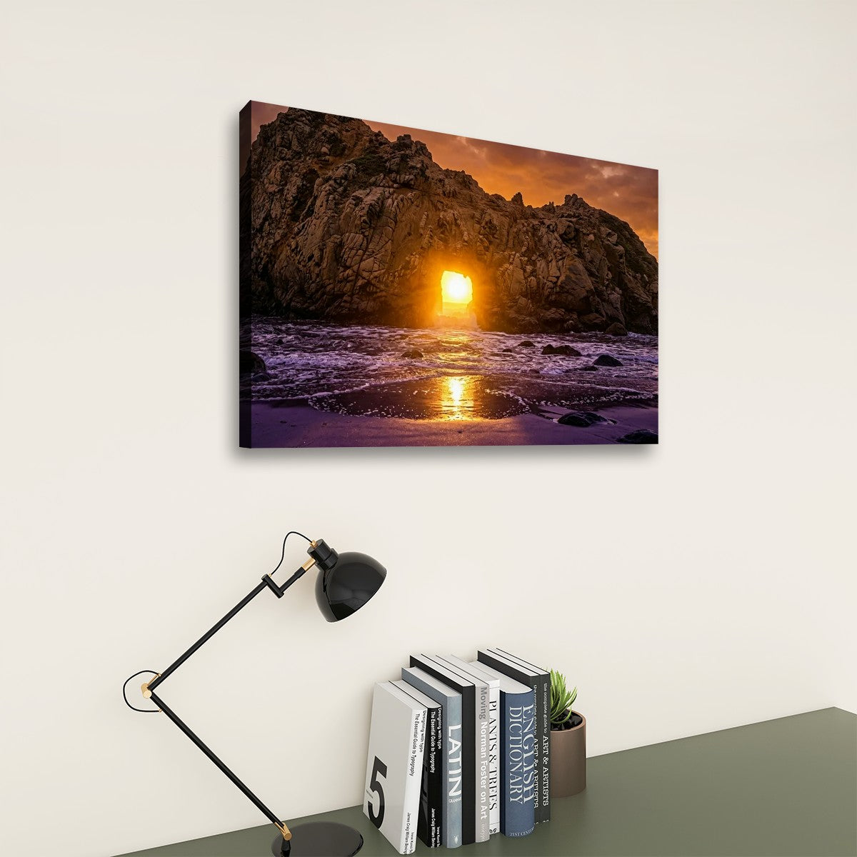 A rectangular wall art print showing a rocky coastline at sunset hangs on a light beige wall above a dark gray table with a black desk lamp and books.