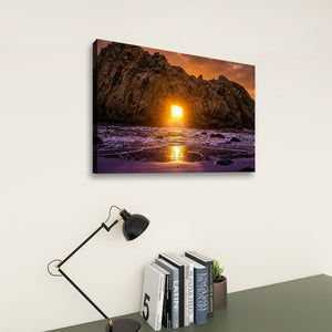 A rectangular wall art print showing a rocky coastline at sunset hangs on a light beige wall above a dark gray table with a black desk lamp and books.