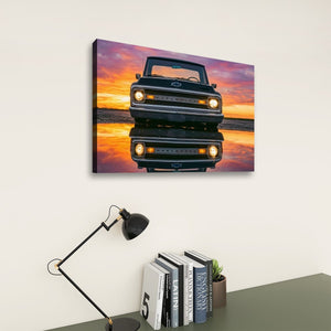 A rectangular wall art print showing a black classic Chevrolet truck reflected in water at sunset, displayed on a light wall above a desk with books, a small plant, and a black desk lamp.