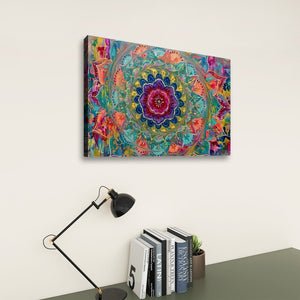 A colorful square wall art print featuring a vibrant mandala design, displayed on a light beige wall above a desk with a black lamp and books.