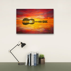 A rectangular wall art print showing a vivid sunset over water with two tree silhouettes reflected in the water, displayed above a desk with a black lamp, books, and a small succulent against a light wall.