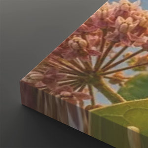 A close-up angled view of a rectangular canvas or print showing a close-up floral scene with pink blossoms, green leaves, and a pale blue sky. The product is photographed against a dark gray studio background.