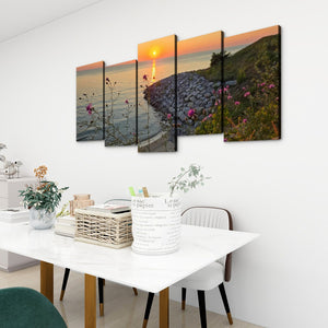 A five-panel canvas wall art set depicting a coastal sunset scene with rocks, water, and pink flowers, displayed above a white dining table in a modern room setting.