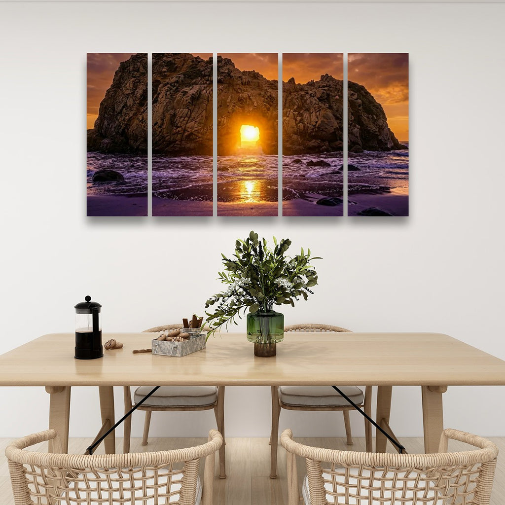 A four-panel canvas wall art set depicting a dramatic rocky coastline at sunset, displayed above a light wood dining table with chairs and decorative items in a modern room setting.