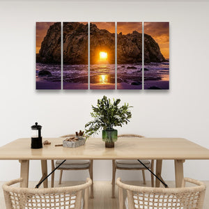 A four-panel canvas wall art set depicting a dramatic rocky coastline at sunset, displayed above a light wood dining table with chairs and decorative items in a modern room setting.