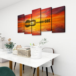 A 5-panel canvas wall art set depicting a guitar silhouette against a vibrant orange and red sunset sky, displayed on a white wall above a dining table in a modern room setting.