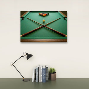 A framed wall art print showing a green pool table with crossed pool cues and a racked set of billiard balls, displayed above a desk with a black lamp, books, and a small succulent on a light wall.