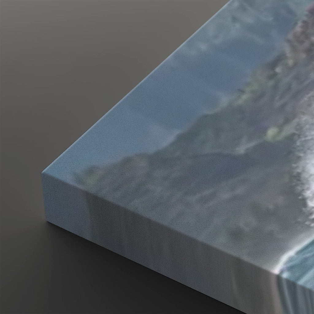 A close-up of a canvas print showing a coastal mountain scene with blue sky and ocean waves, photographed at an angle on a dark gray background.