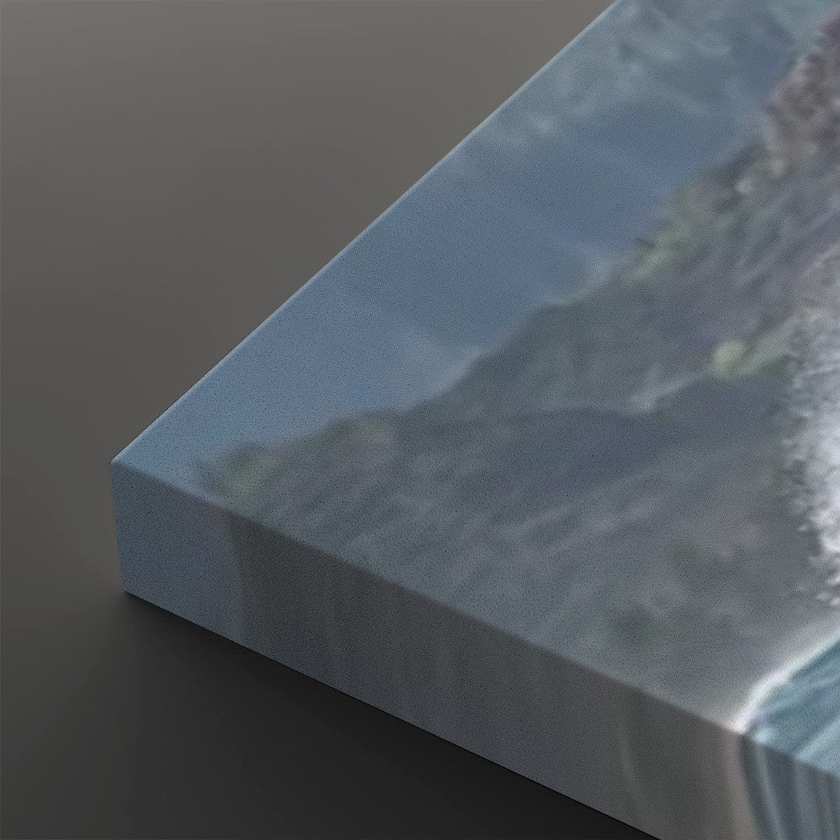 A close-up of a canvas print showing a coastal mountain scene with blue sky and ocean waves, photographed at an angle on a dark gray background.
