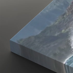 A close-up of a canvas print showing a coastal mountain scene with blue sky and ocean waves, photographed at an angle on a dark gray background.
