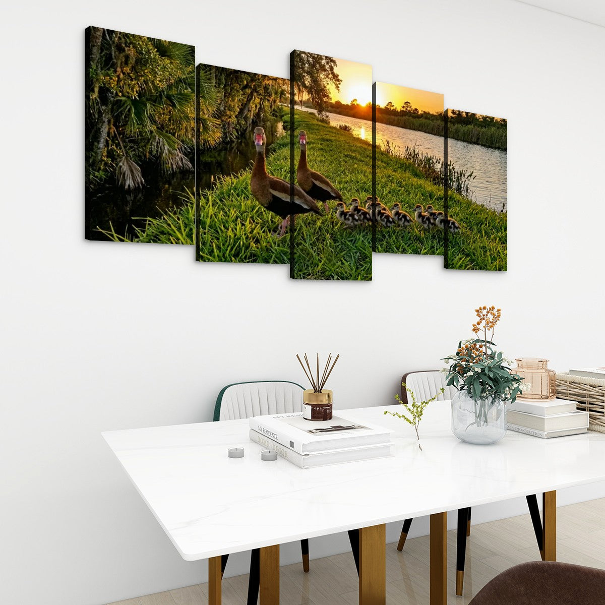 A five-panel canvas wall art set depicting a mother duck and her ducklings by a lake at sunset, displayed on a white wall above a modern dining table with chairs and decorative items.