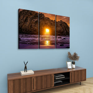 A three-panel canvas wall art set depicting a rocky coastline at sunset over the ocean, displayed on a light blue wall above a wooden console table with decorative items.