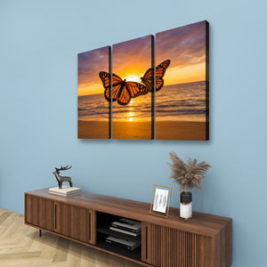 Three-panel canvas wall art showing a butterfly over a beach sunset, displayed above a wooden console in a styled room with a blue wall.