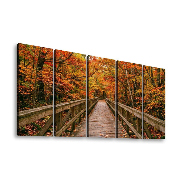 A five-panel canvas wall art set depicting a wooden boardwalk bridge spanning through an autumn forest with vibrant orange, red, and yellow foliage.