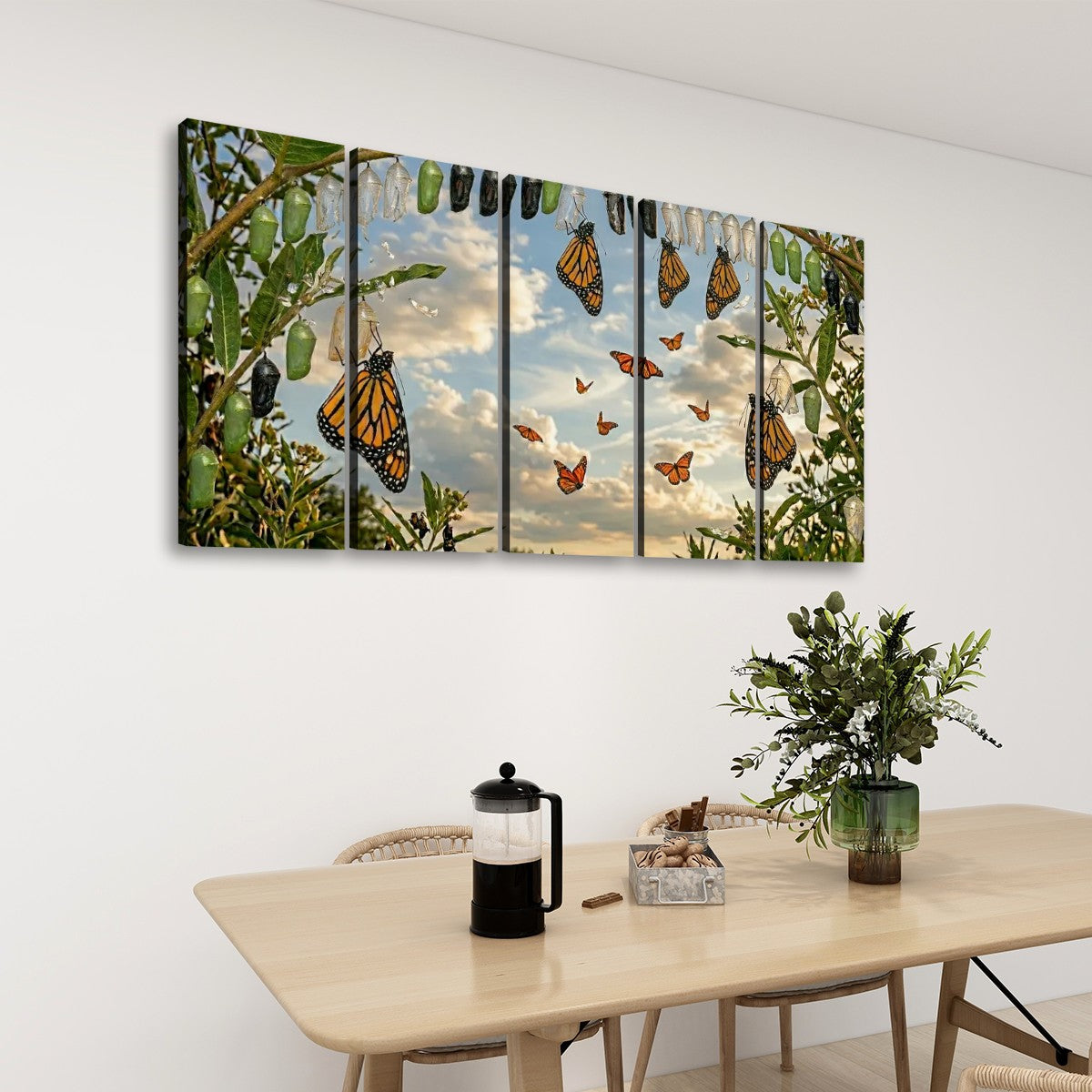 A five-panel canvas wall art set depicting monarch butterflies in a garden under a cloudy sky, displayed on a white wall above a wooden dining table with a vase of greenery and a black coffee press.