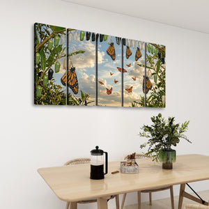 A five-panel canvas wall art set depicting monarch butterflies in a garden under a cloudy sky, displayed on a white wall above a wooden dining table with a vase of greenery and a black coffee press.