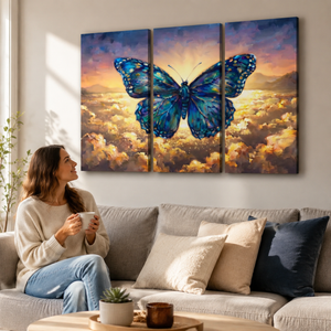 A woman sits on a light gray sofa holding a white mug, looking at a three-panel canvas wall art set depicting a large blue butterfly over a glowing golden sky and clouds.