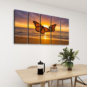 A five-panel canvas wall art set depicting two orange monarch butterflies over a sunset beach scene, displayed on a white wall above a dining table with a plant and coffee press.