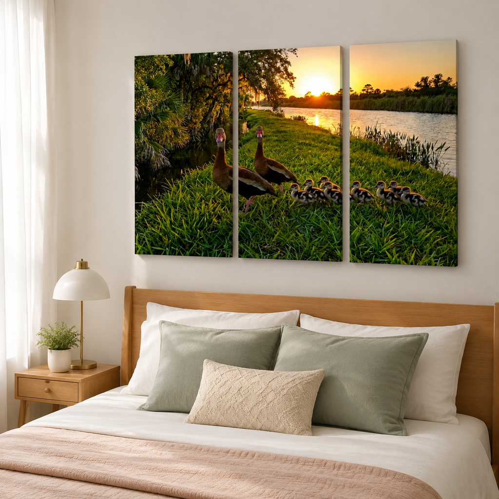 A three-panel canvas wall art print depicting a mother duck and her ducklings by a lake at sunset, displayed above a bed in a modern bedroom setting.