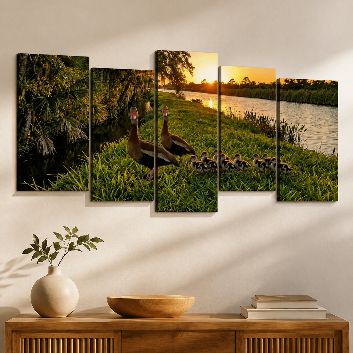 A five-panel canvas wall art set depicting a mother duck and her ducklings by a lake at sunset, displayed on a wall above a wooden console table with a vase and books.