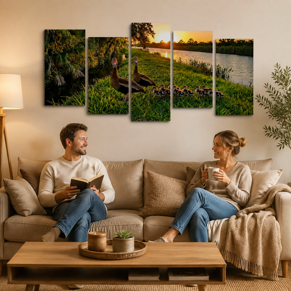 A five-panel canvas wall art set depicting a family of Black-bellied Whistling-Ducks and ducklings by a lake at sunset, displayed on a beige wall above a living room sofa where a man and woman are sitting and reading.