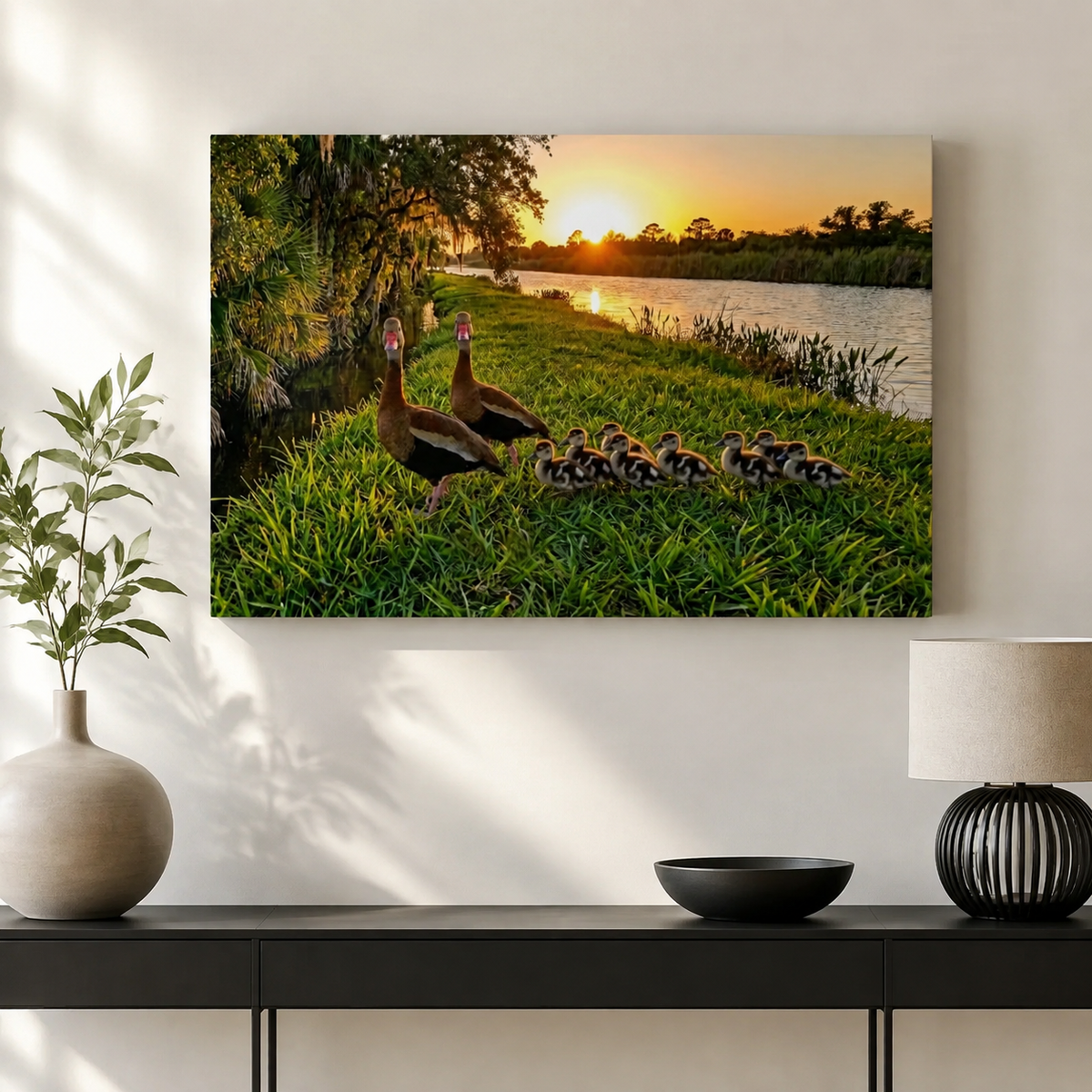 A rectangular wall art print showing two adult Black-bellied Whistling-Ducks and several ducklings on a grassy lakeside at sunset, displayed above a console table in a styled interior with a vase, bowl, and lamp.