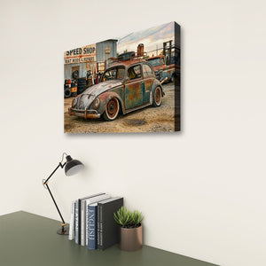 A rectangular wall art print showing a vintage rusted Volkswagen Beetle in an industrial workshop scene, displayed above a desk with books, a small plant, and a black desk lamp against a light wall.