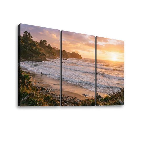 Three-panel canvas wall art showing a coastal beach and ocean at sunset, with cliffs, waves, sand, and golden sky tones, displayed against a light gray background.