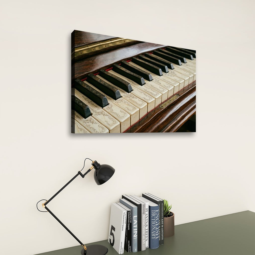 A rectangular wall art print showing a close-up of a piano keyboard hangs on a light beige wall above a desk with a black lamp and books.