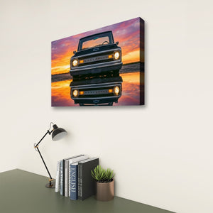 A rectangular wall art print showing a red vintage 1970 Chevy C10 reflected in water at sunset, displayed on a light wall above a desk with a lamp, books, and a small plant.