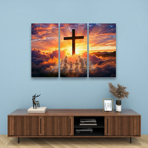 A three-panel canvas wall art set depicting a silhouette of a Christian cross over a dramatic sunset sky with clouds and angelic figures, displayed on a light blue wall above a wooden console table with decorative items.