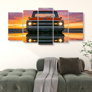 A 4-panel canvas wall art print depicting a classic orange Chevrolet truck at sunset, displayed on a white wall above a green tufted sofa in a living room setting.