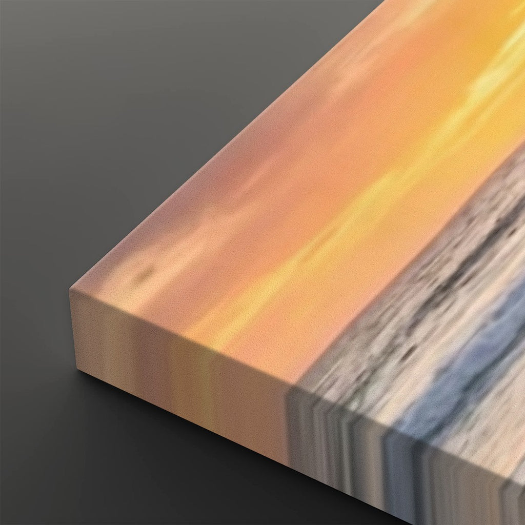A close-up of a rectangular art print or canvas showing a sunset seascape with warm orange and yellow sky, pale sand, and blue-gray water, photographed on a dark gray background.