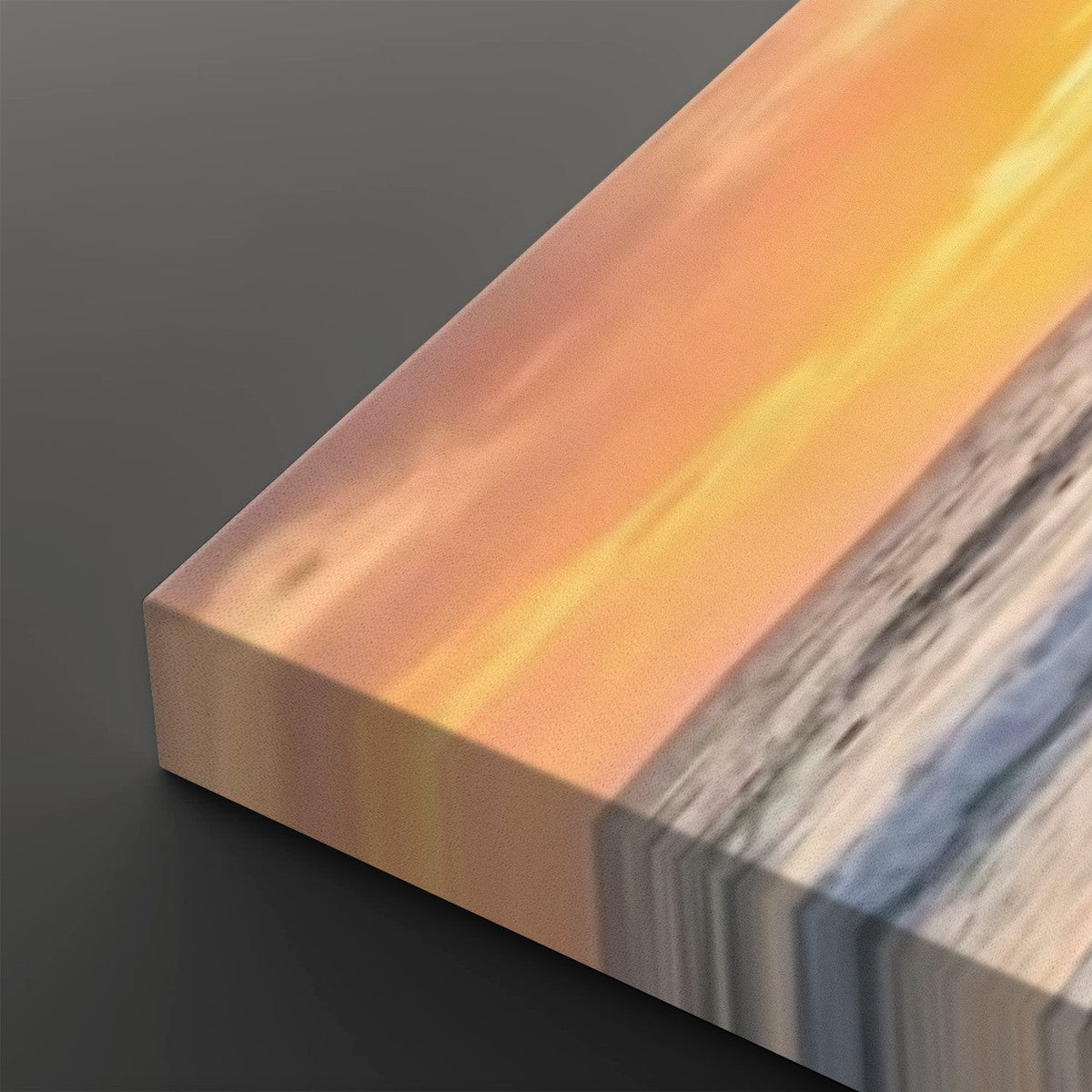 A close-up of a rectangular art print or canvas showing a sunset seascape with warm orange and yellow sky, pale sand, and blue-gray water, photographed on a dark gray background.