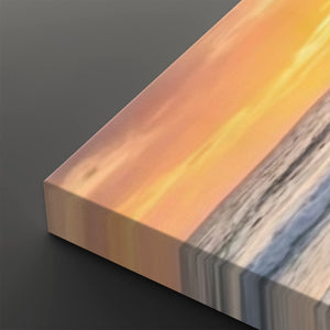 A close-up of a rectangular art print or canvas showing a sunset seascape with warm orange and yellow sky, pale sand, and blue-gray water, photographed on a dark gray background.
