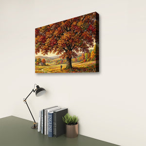 A rectangular wall art print showing a large autumn tree with orange and yellow foliage over a rolling landscape, displayed on a light wall above a desk with books, a lamp, and a small plant.