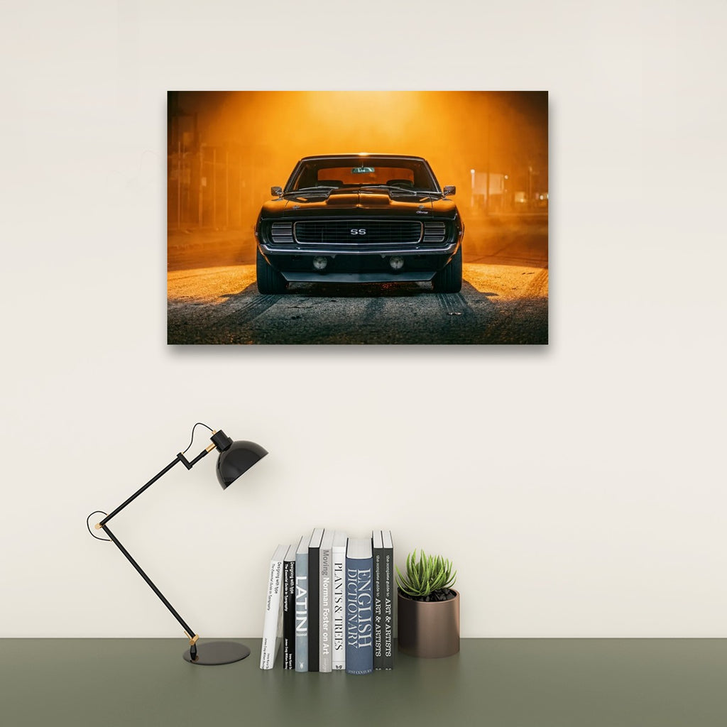 A framed wall art print showing a black muscle car facing forward against a dramatic orange-yellow smoky background, displayed above a desk with a black lamp, books, and a small potted succulent.