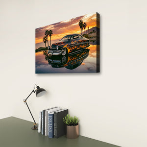 A rectangular wall art print showing a black classic car with orange flame decals reflected in water, hung on a light wall above a desk with a lamp, books, and a small plant.