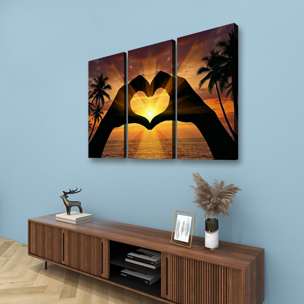 Three-panel canvas wall art showing a sunset seascape with silhouetted hands forming a heart shape, displayed above a wooden console in a styled room with a blue wall.