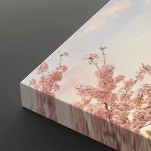 A rectangular wall art canvas showing a soft-focus pink cherry blossom scene against a pale sky, photographed at an angle on a dark gray background.
